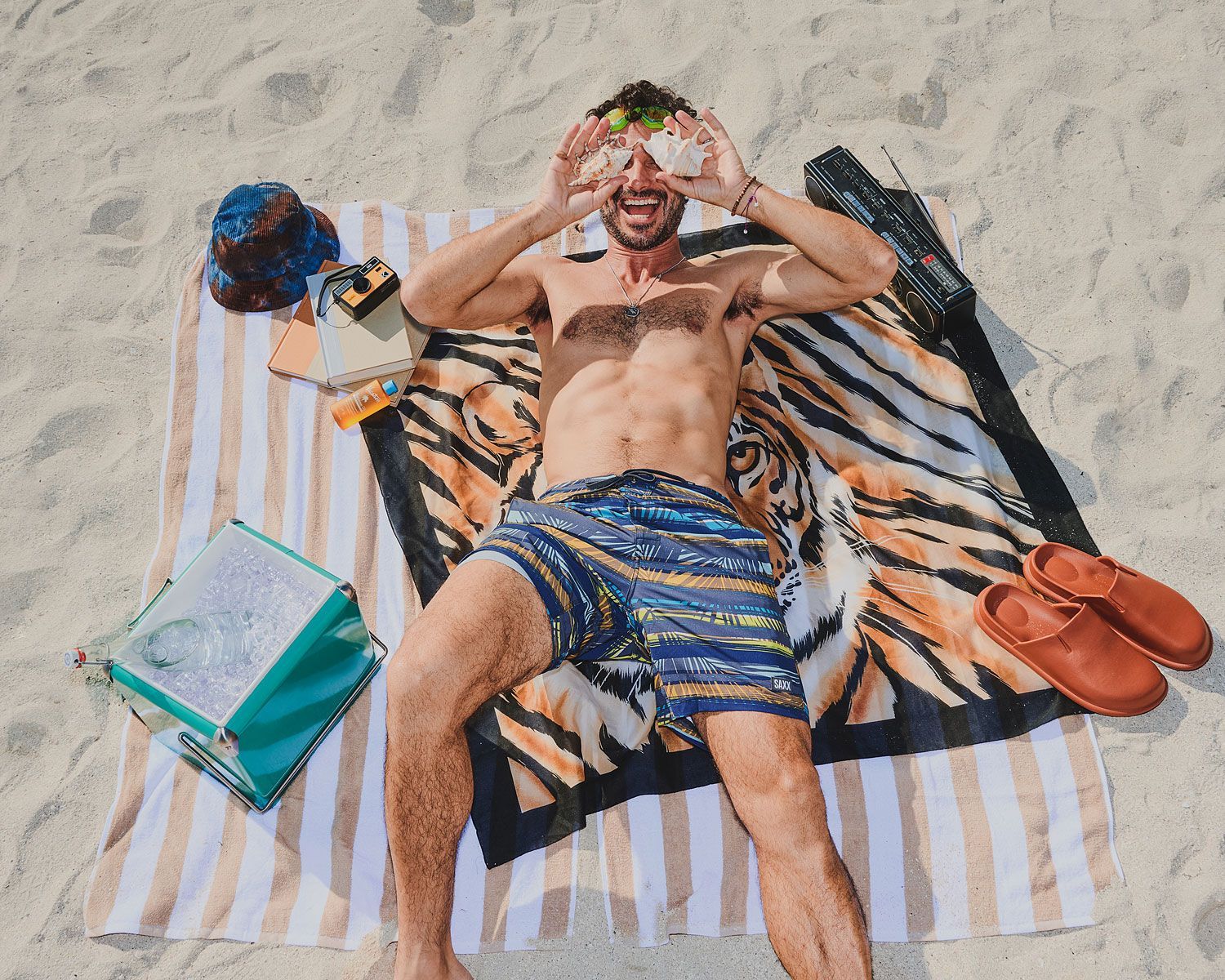 Man on beach holding food over eyes; lying on towel, smiling.
