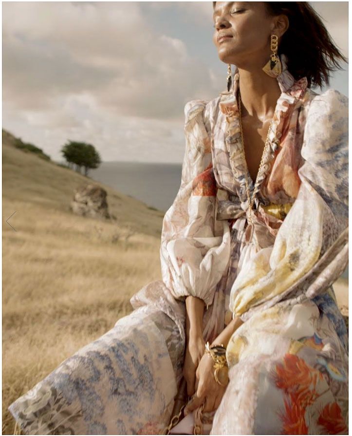 Woman in patterned dress sits on a grassy hill overlooking the sea, eyes closed, sunshine.
