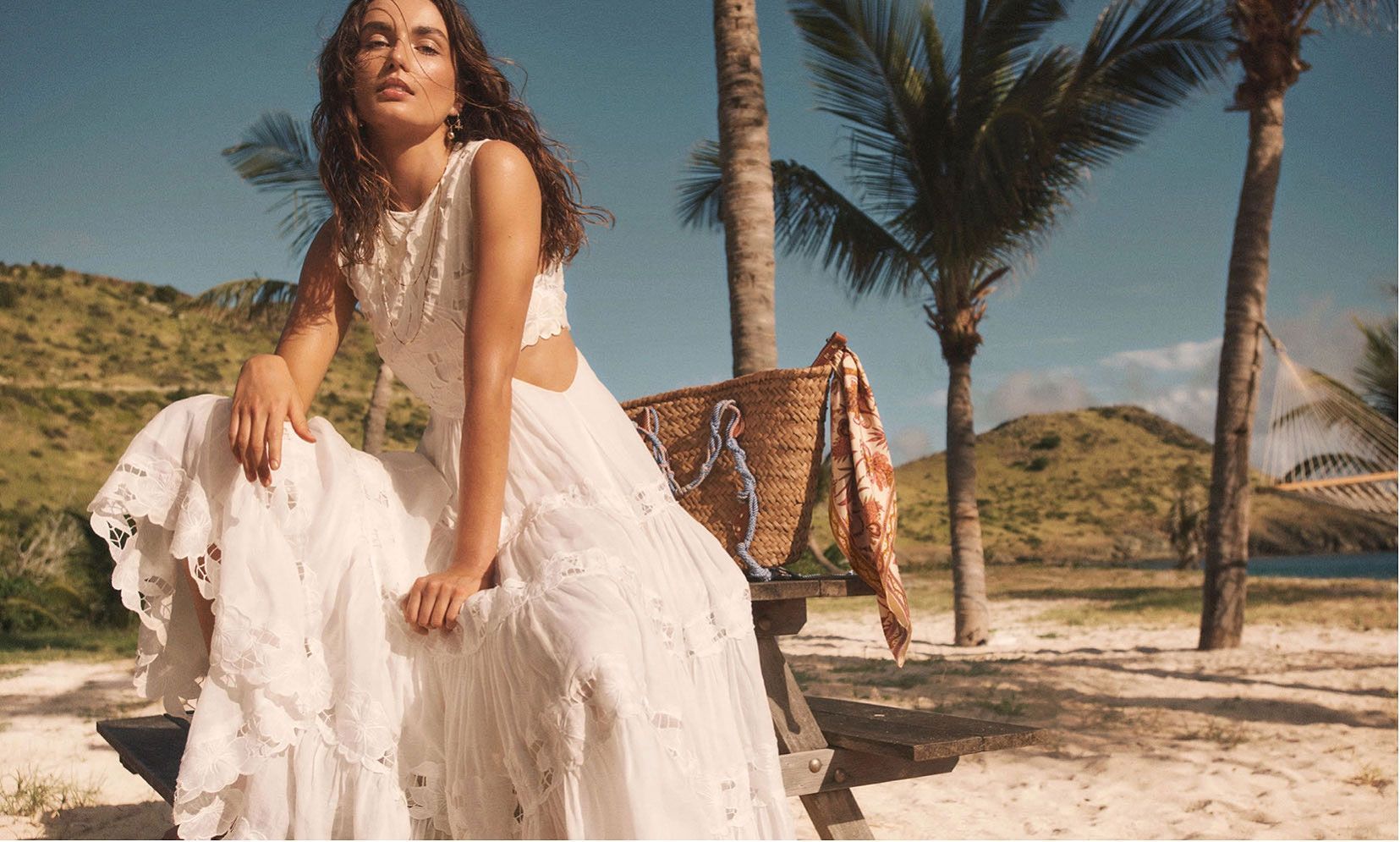 Woman in white dress sits on a bench on a beach, posing in the sun.