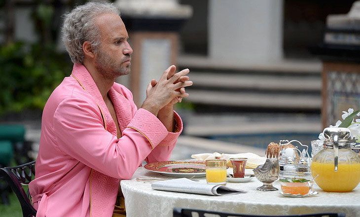 Man in pink robe sits at outdoor table, hands clasped.  Breakfast setting with orange juice, near a pool.