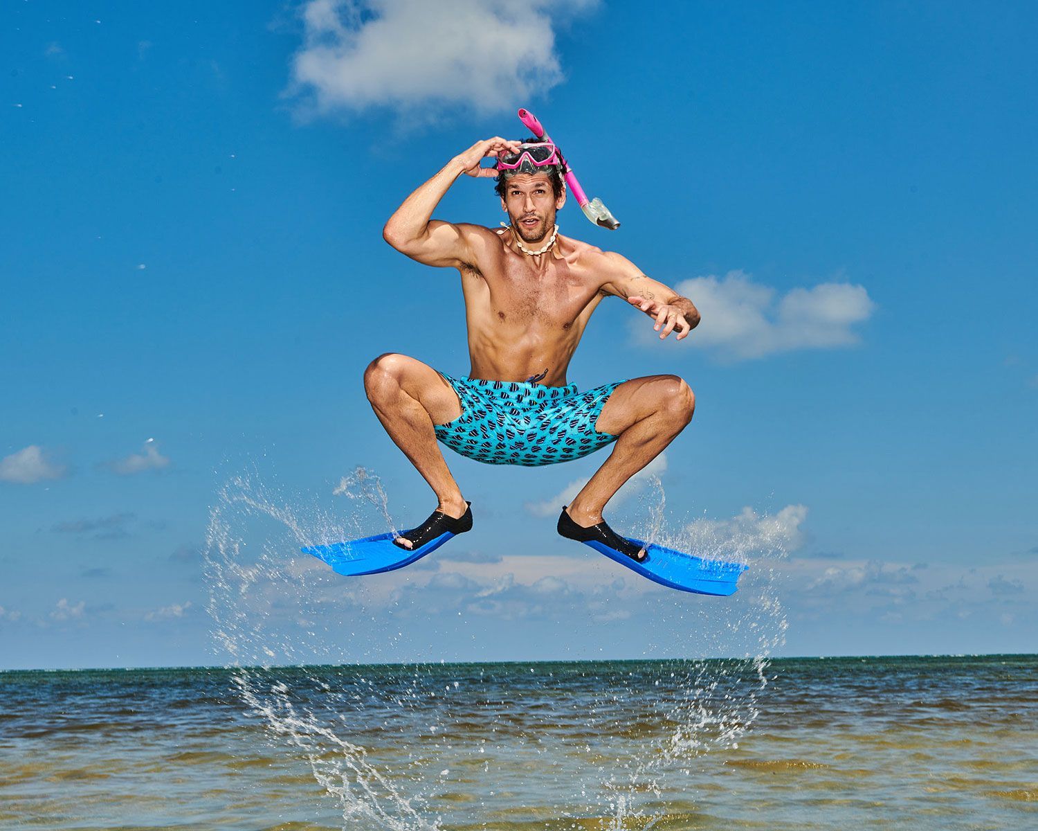 Man in blue swim trunks and flippers jumps in the air, wearing snorkel gear. Water splashes. Sunny beach.