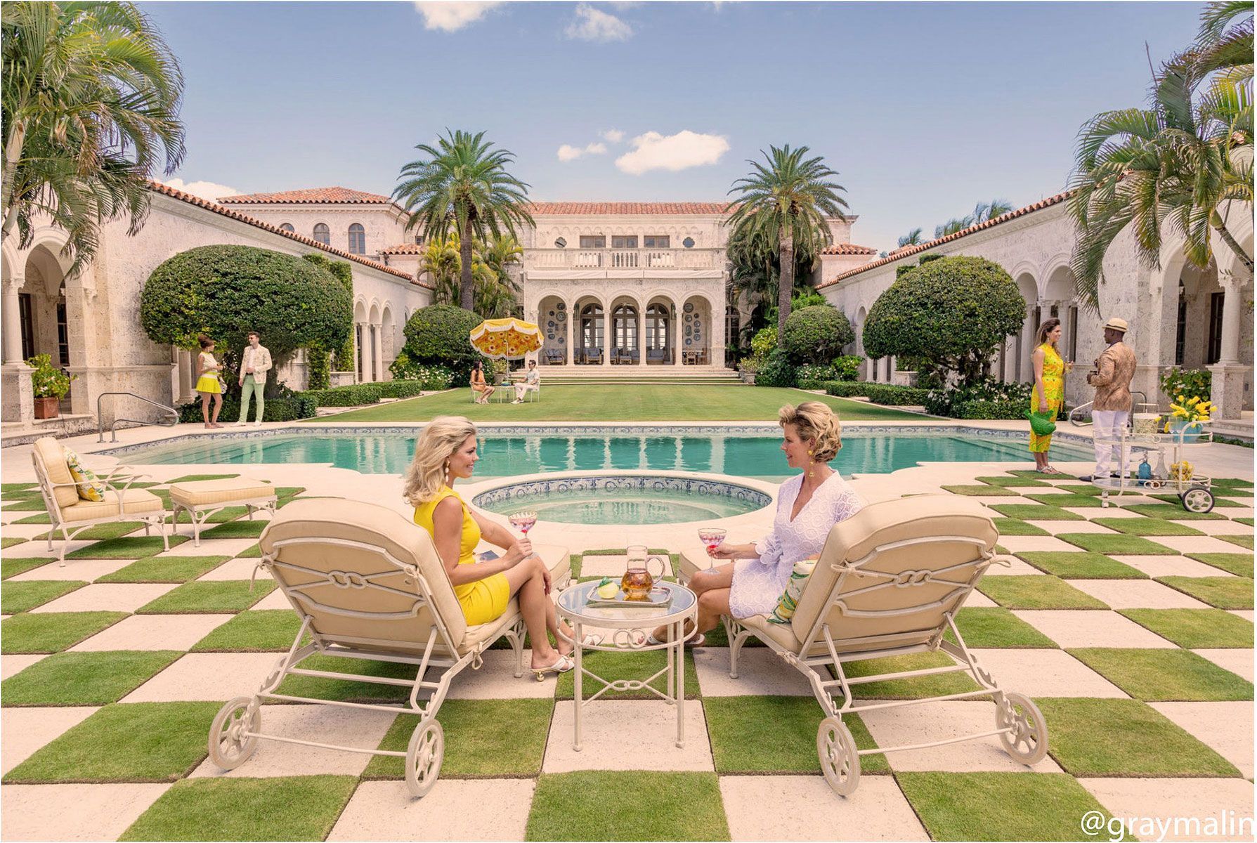 Two women in lounge chairs near a pool at a lavish estate, palm trees and architecture visible.