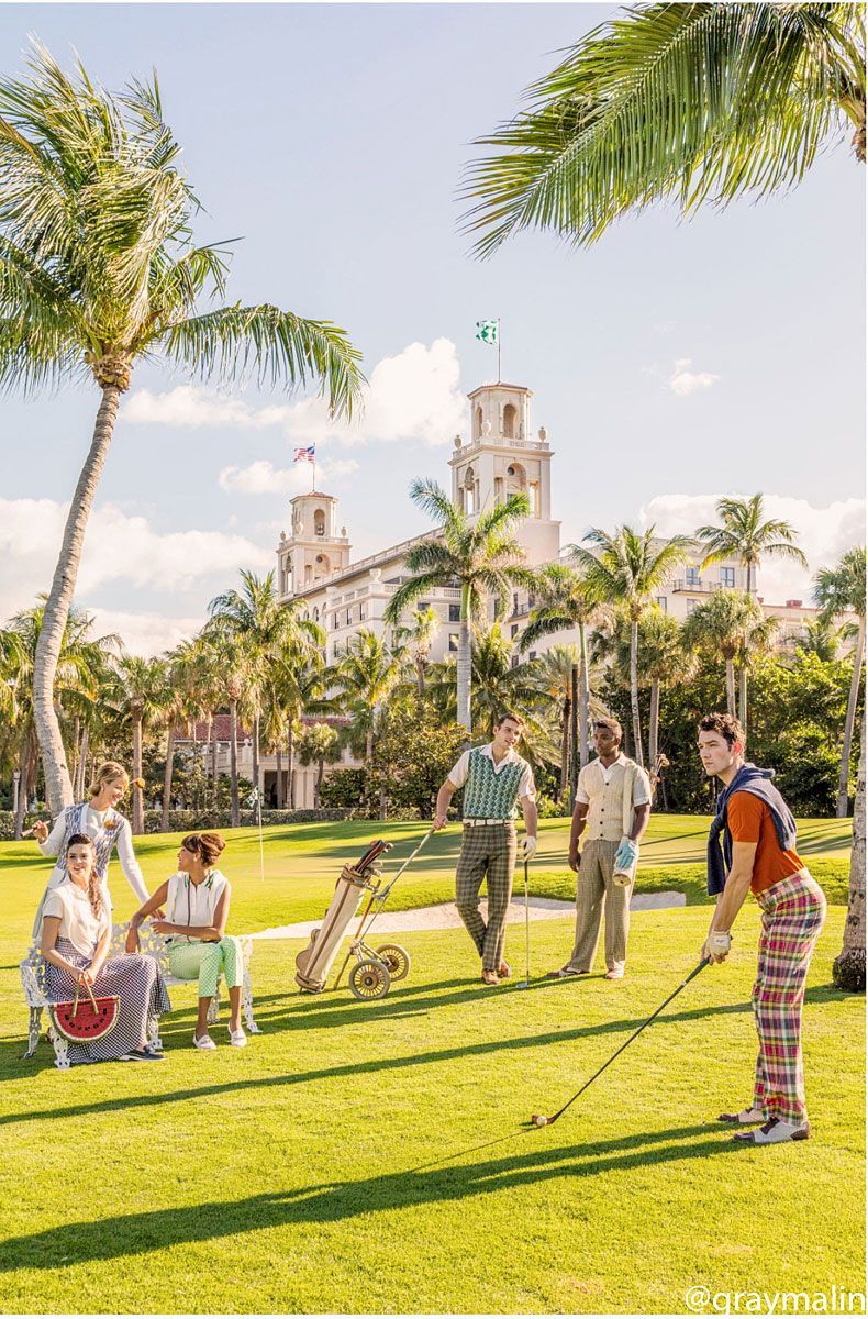People golfing on a green lawn with palm trees and a large building in the background. Bright sunny day.