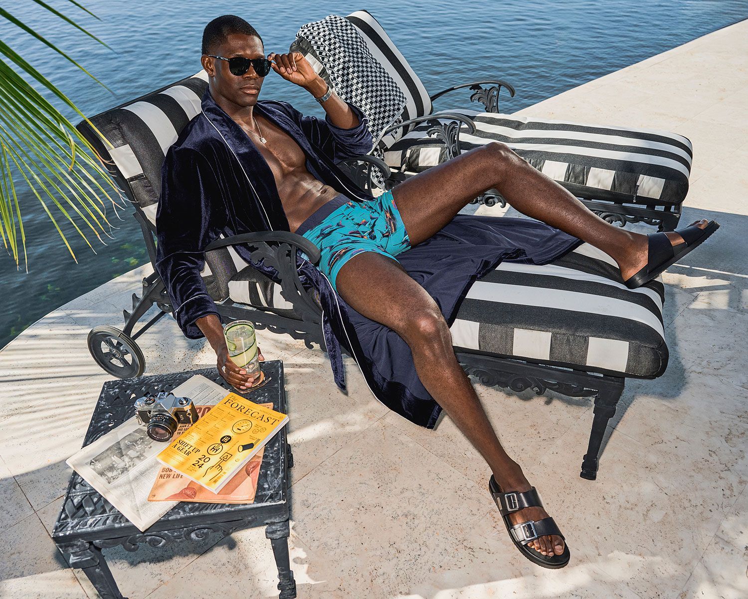 Man in robe and swim trunks relaxes by water, sitting on striped chair.