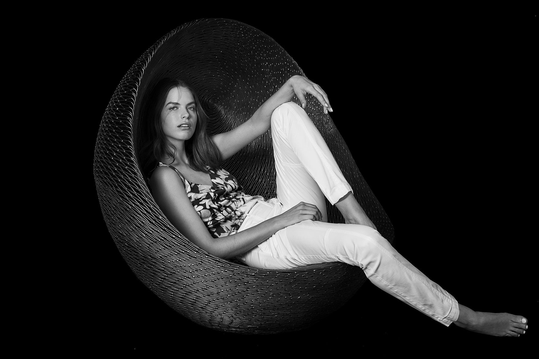Woman lounging in a woven egg chair, wearing floral top and white pants, black and white.