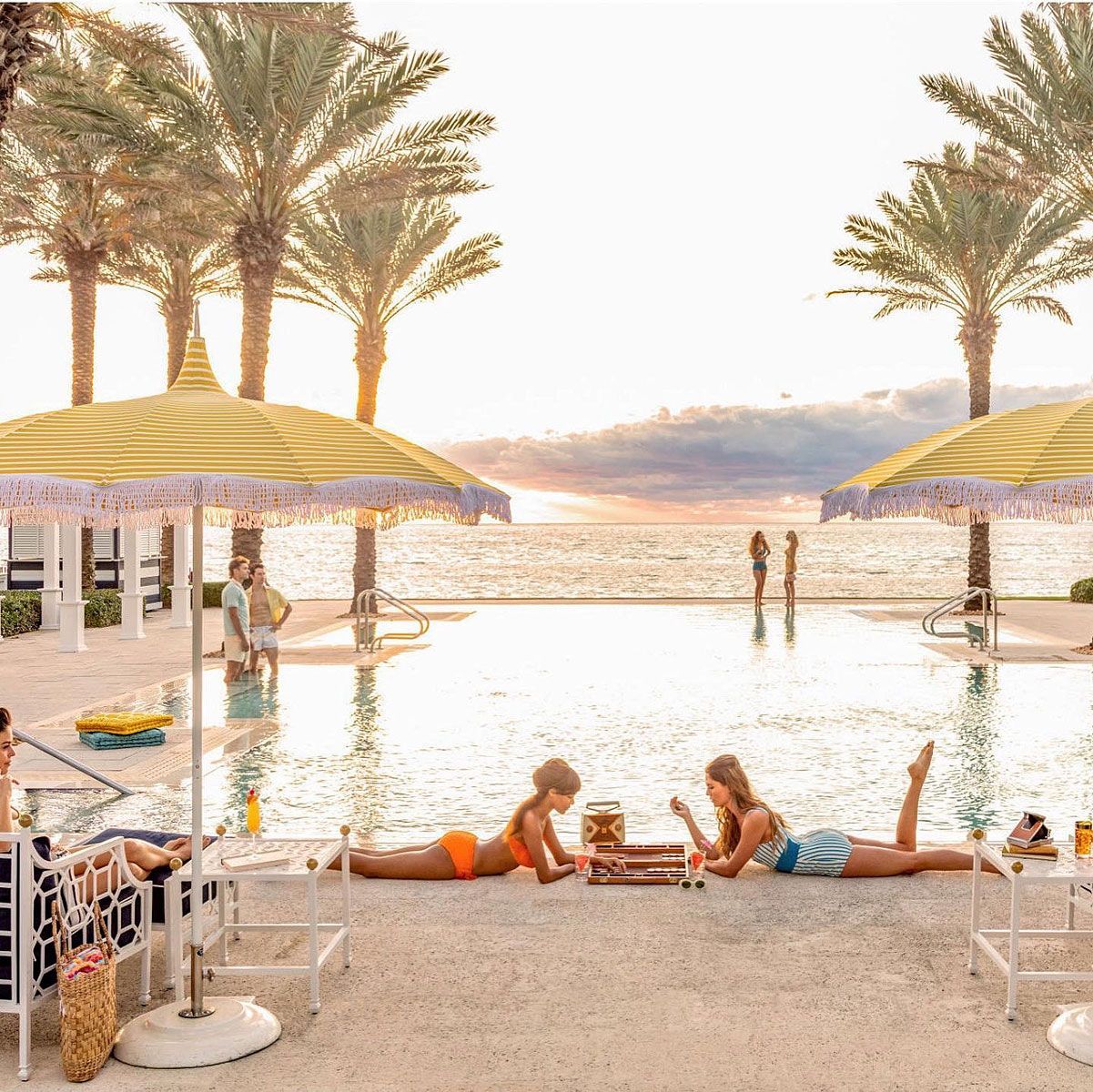 Two women sunbathe by a pool. Yellow umbrellas, palm trees, and ocean in the background.