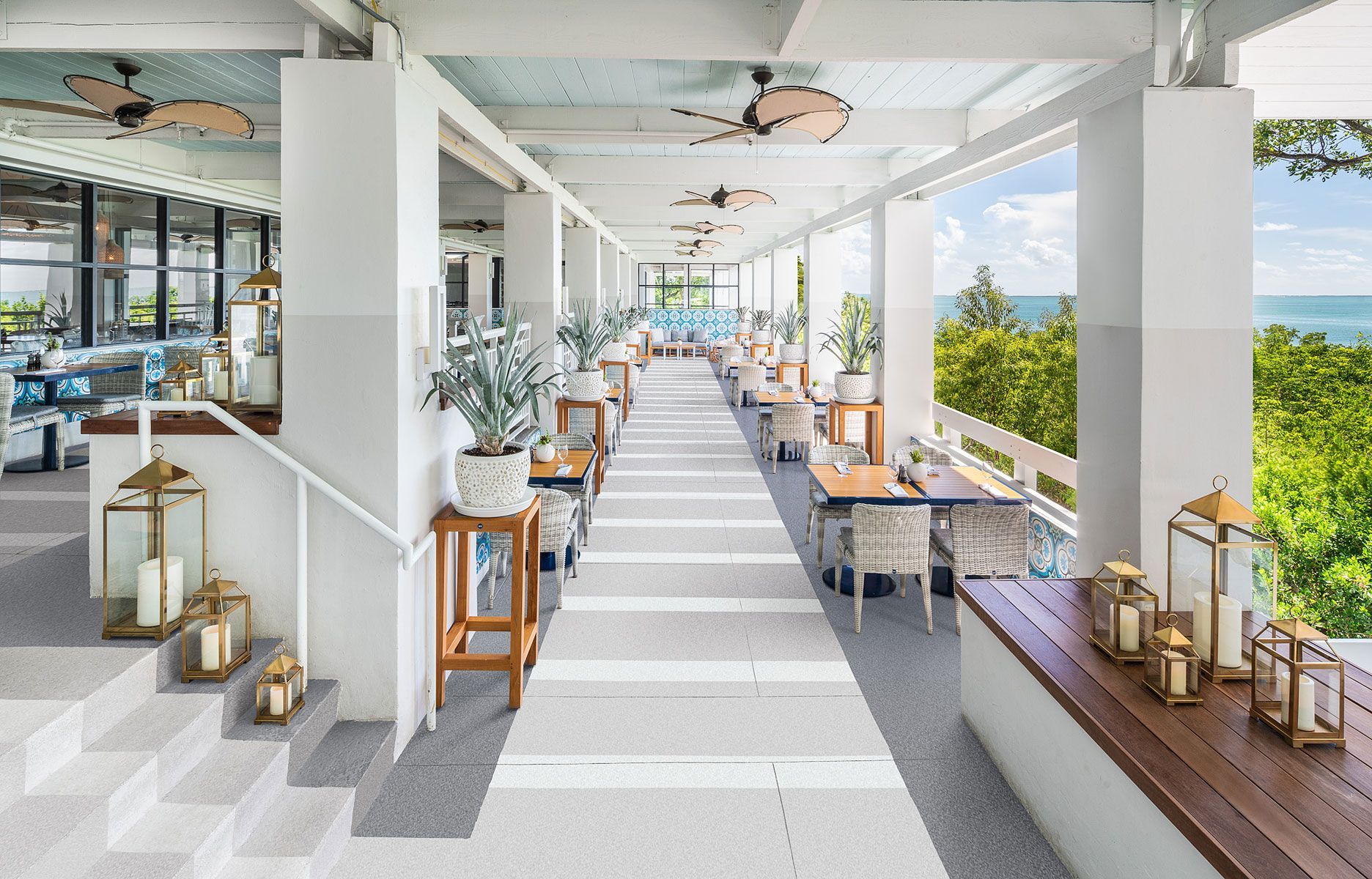 A bright, open-air restaurant patio with ocean views, tables, and lanterns.