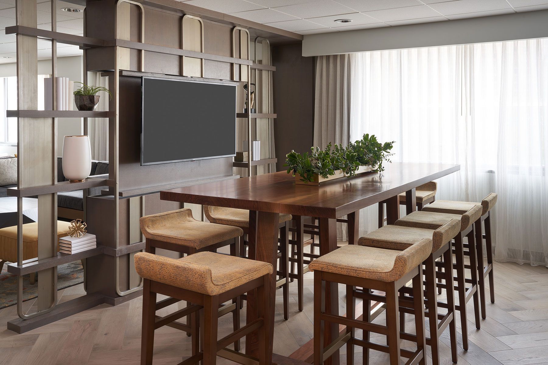 Modern lounge with large wooden table and bar stools, TV, and shelves.