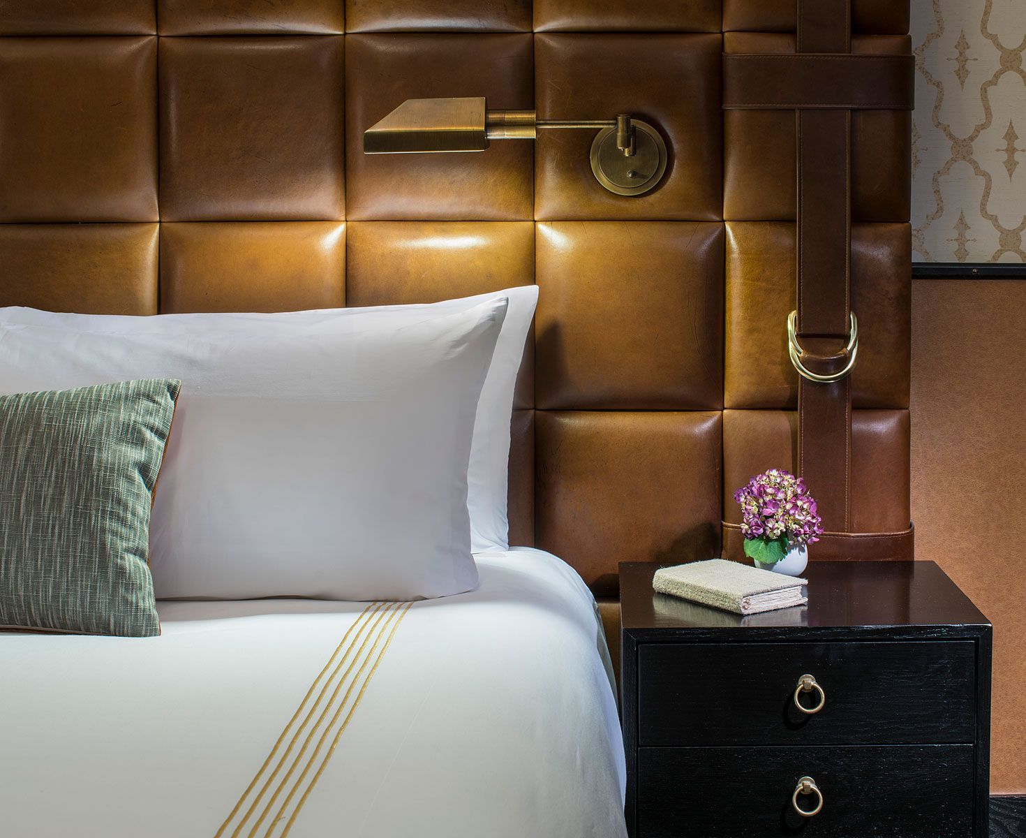 A bed with white linens, a brown leather headboard, a nightstand, and a wall-mounted reading lamp.