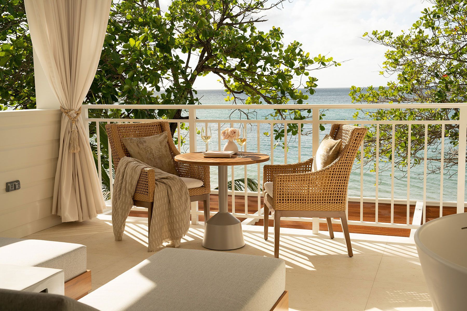 Balcony with woven chairs, small table, ocean view. Light-colored curtains, neutral tones, and foliage.