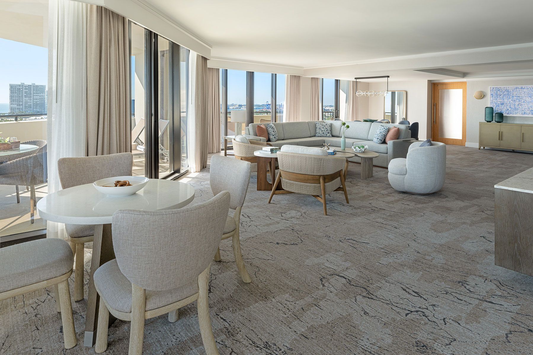 Spacious living room with large windows, neutral tones, and seating areas; dining table in the foreground.