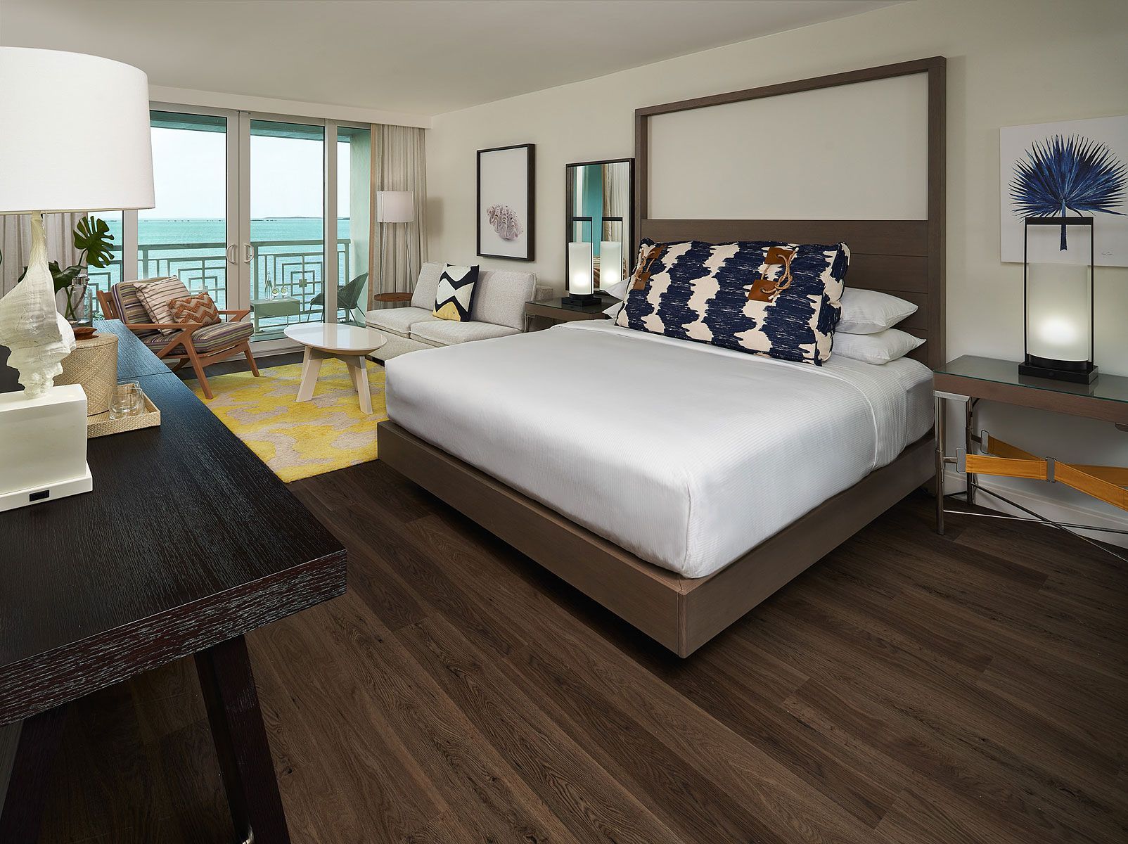 Hotel room with king bed, balcony, and ocean view. Dark wood floor, neutral walls, and colorful accents.