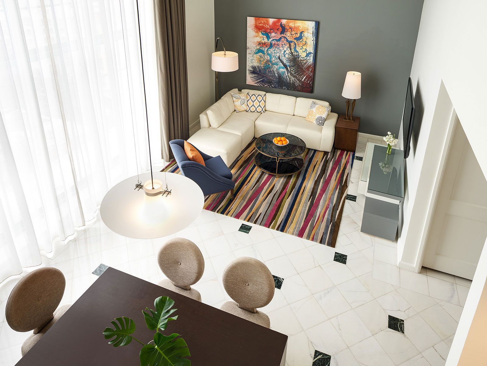 High-angle view of a modern living room. Features sectional sofa, dining table, rug, artwork, and large windows.
