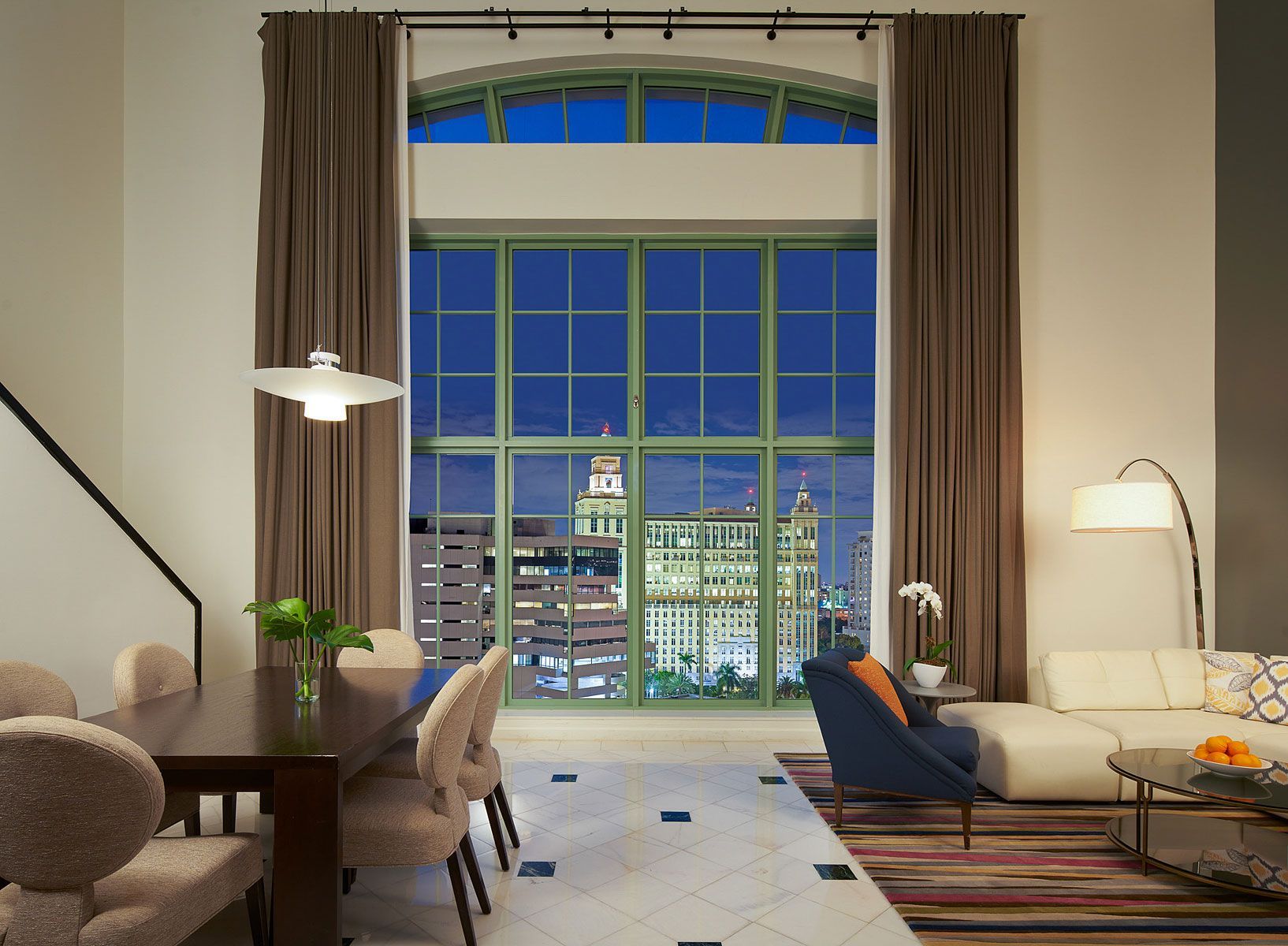 Living room with large windows, city view at night, dining table, sofa, and accent chair.