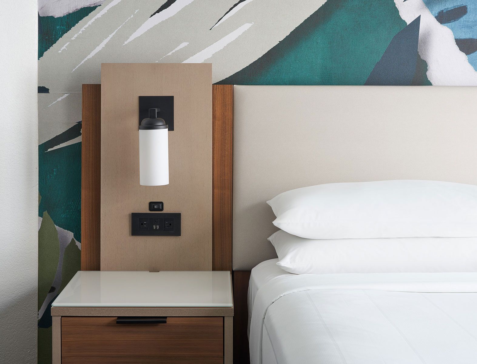 Hotel room bed with nightstand, lamp, and outlet. Neutral colors, modern design, decorative wall art in background.