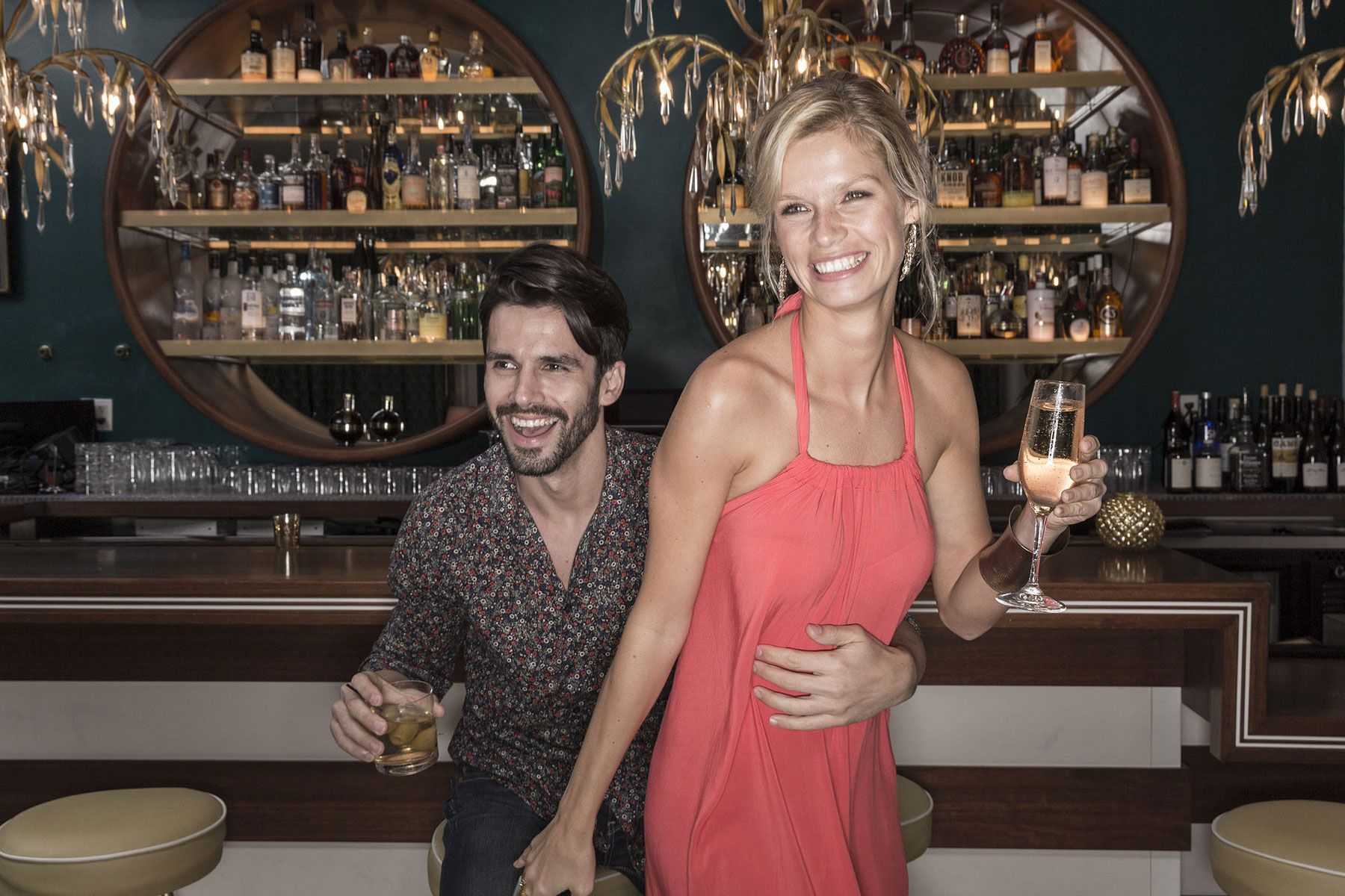 Man and woman at a bar, smiling. Woman in red dress holds a drink; man with a drink leans in.