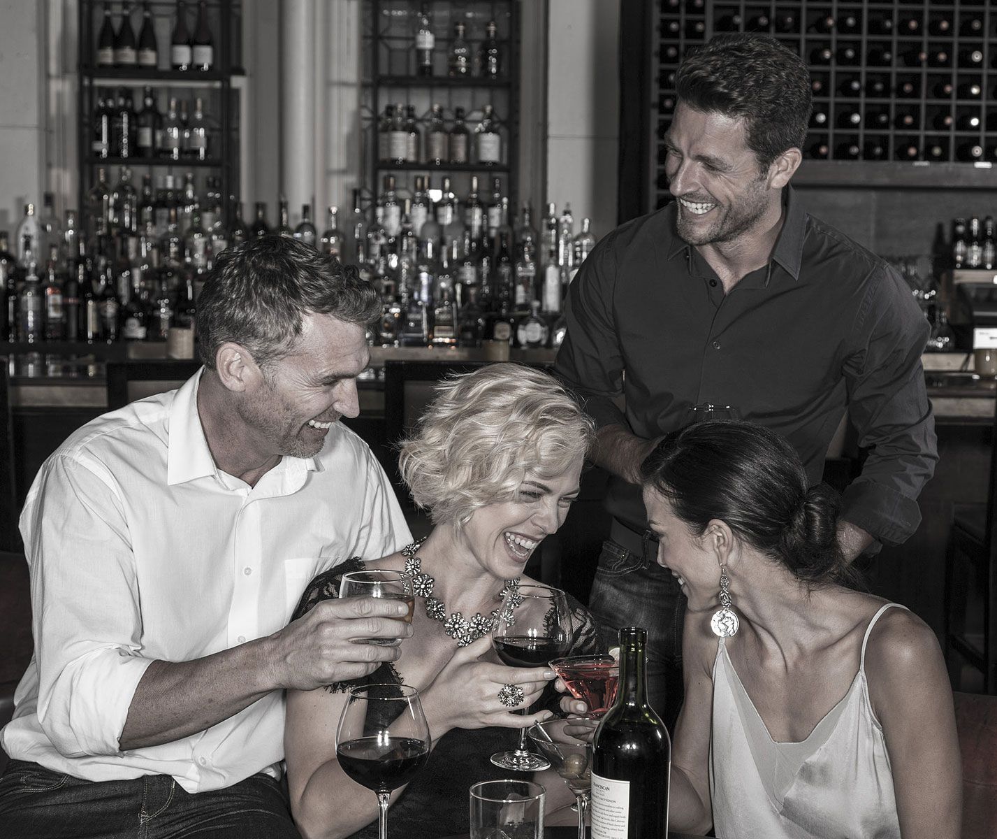 Four people at a bar laughing while holding glasses of wine; a bar with bottles in the background.