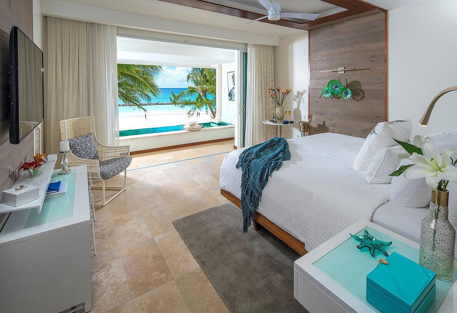 Bedroom with ocean view; white bed, sliding doors open to turquoise water, neutral tones.