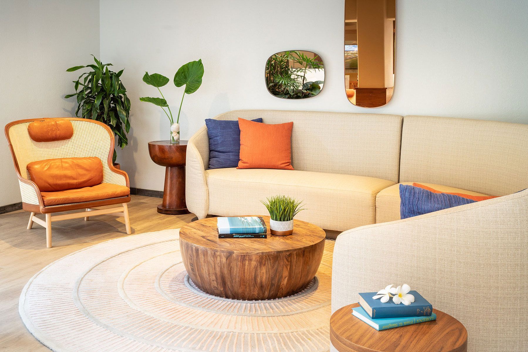 Living room with cream-colored couch, rattan chair, round wooden coffee table, and plants. Blue, orange, and tan decor.