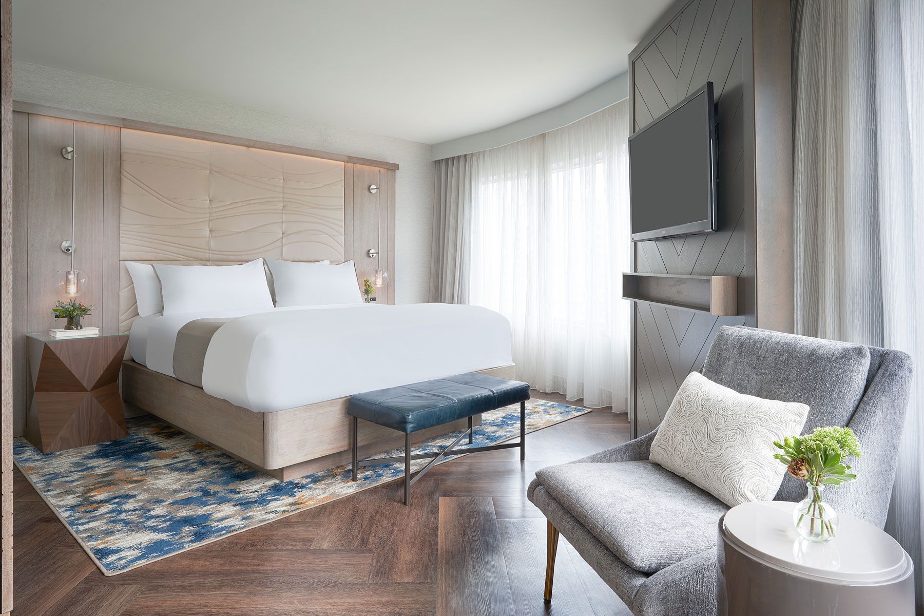 Hotel bedroom with bed, armchair, TV, rug, and side table with flower.
