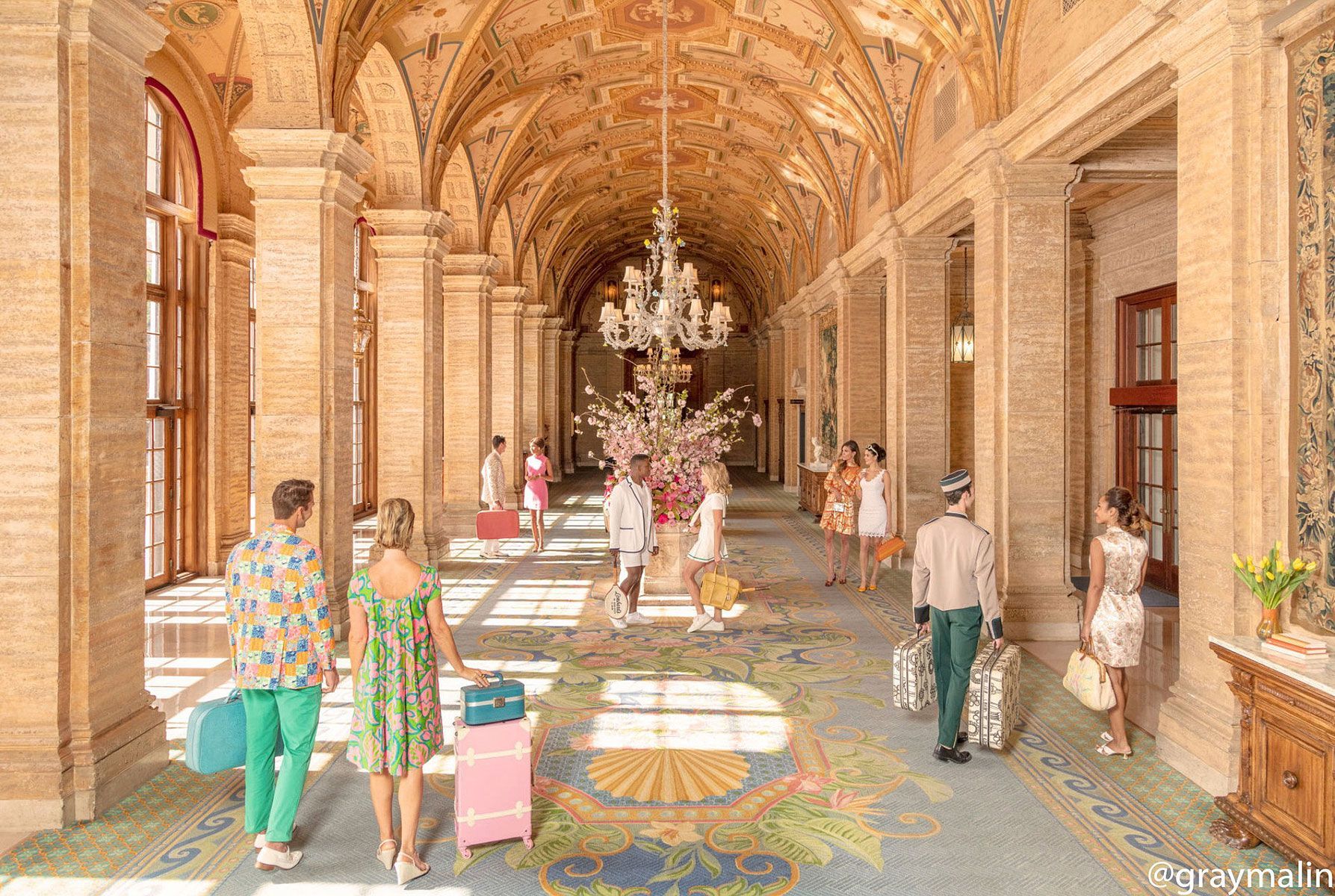 People with luggage walk in a grand, ornate hallway with a chandelier.