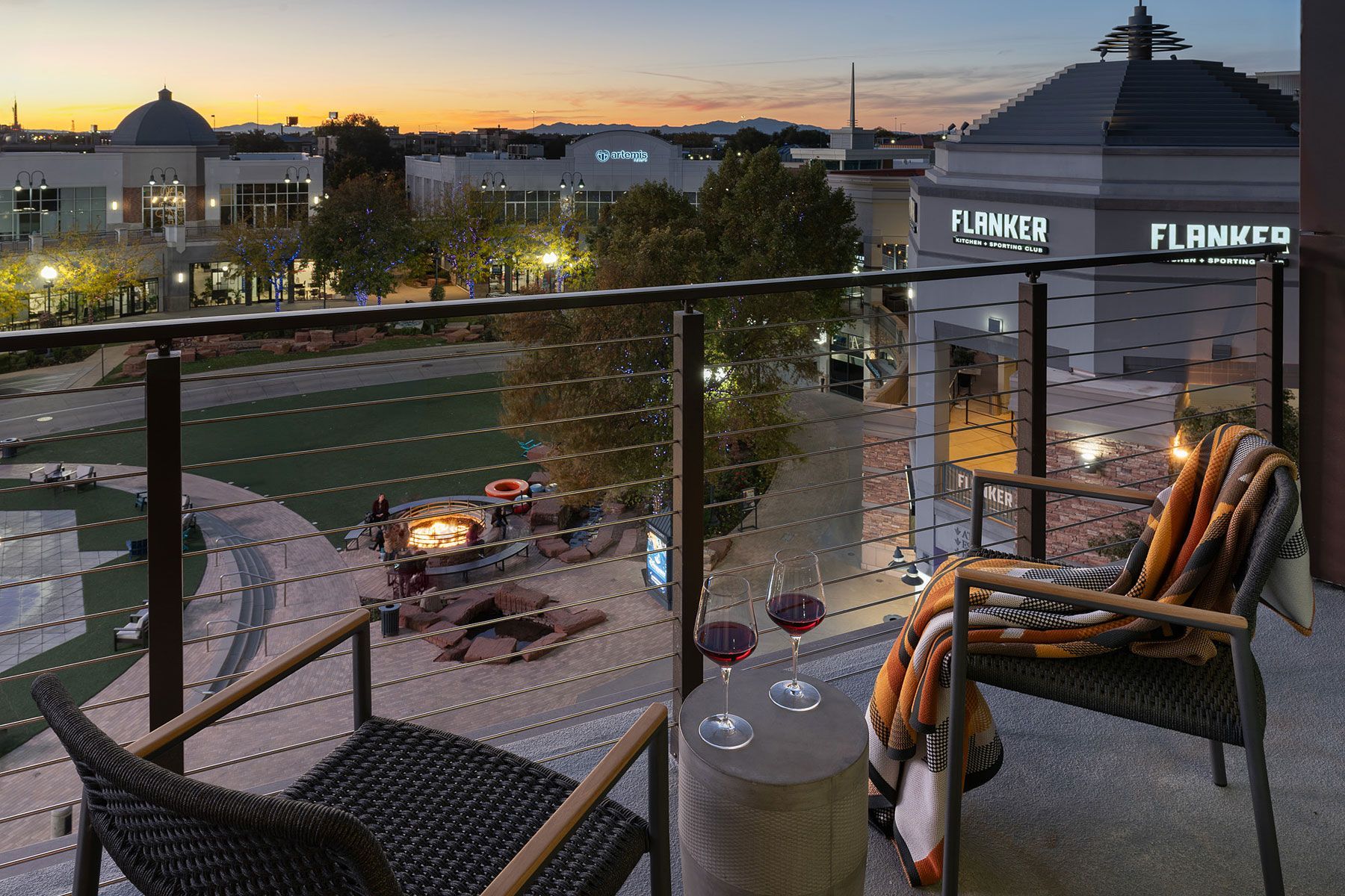 Balcony with two chairs, wine glasses, and a blanket overlooking a town square at dusk.