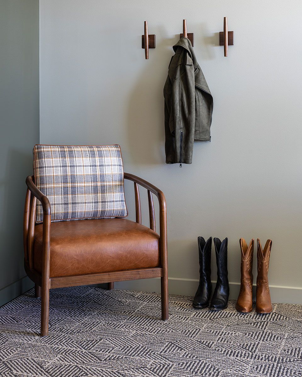 Chair, boots, and coat in a corner. Brown leather chair, olive jacket, and dark boots on patterned rug.