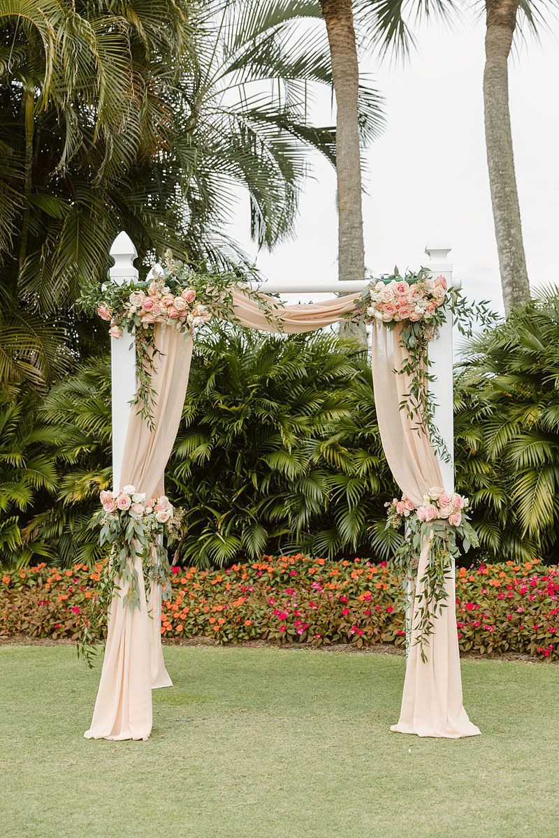 Wedding arch with blush drapes, floral arrangements, and greenery in a garden setting.