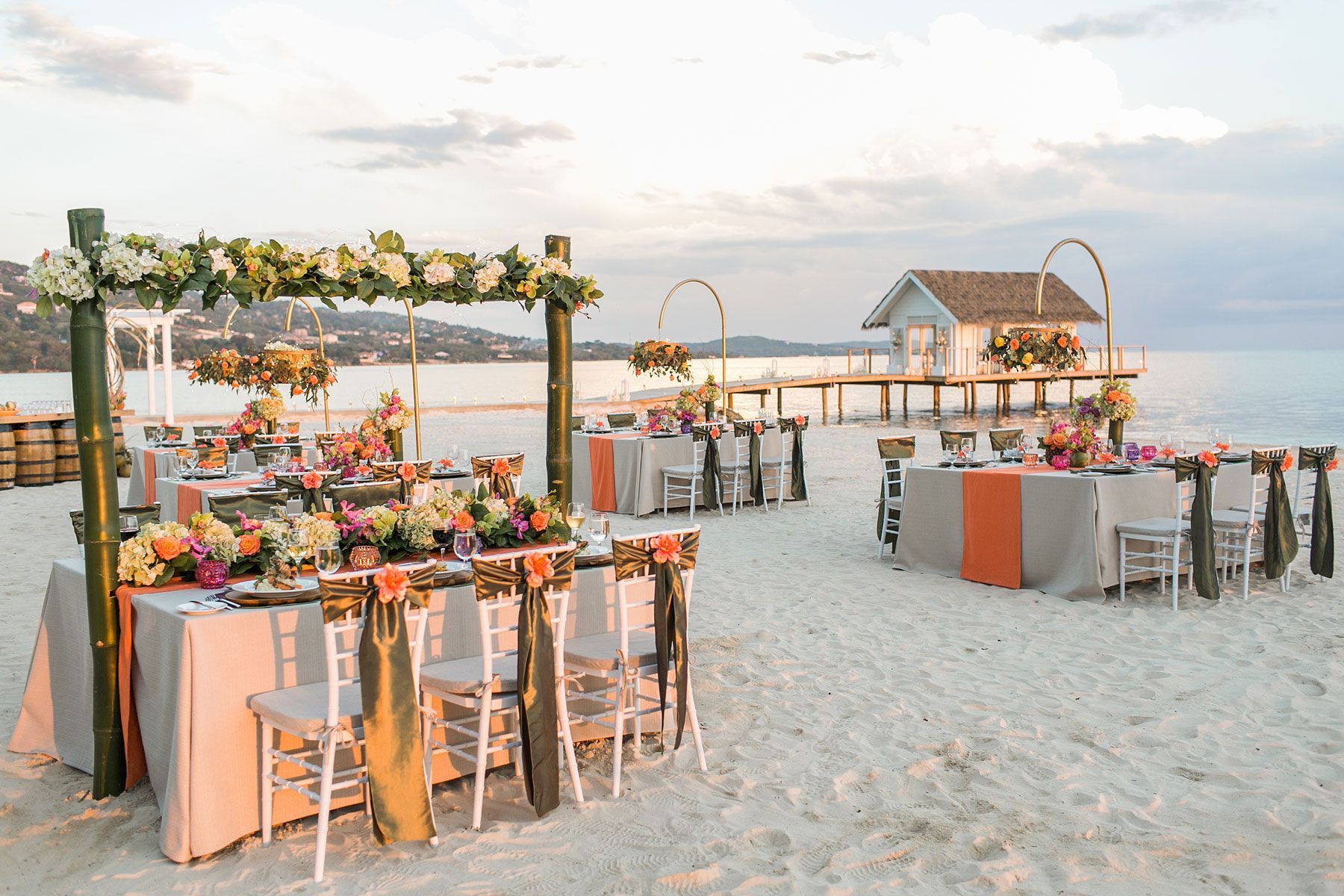 Beach wedding reception with tables set for guests; floral arrangements, sunset background.