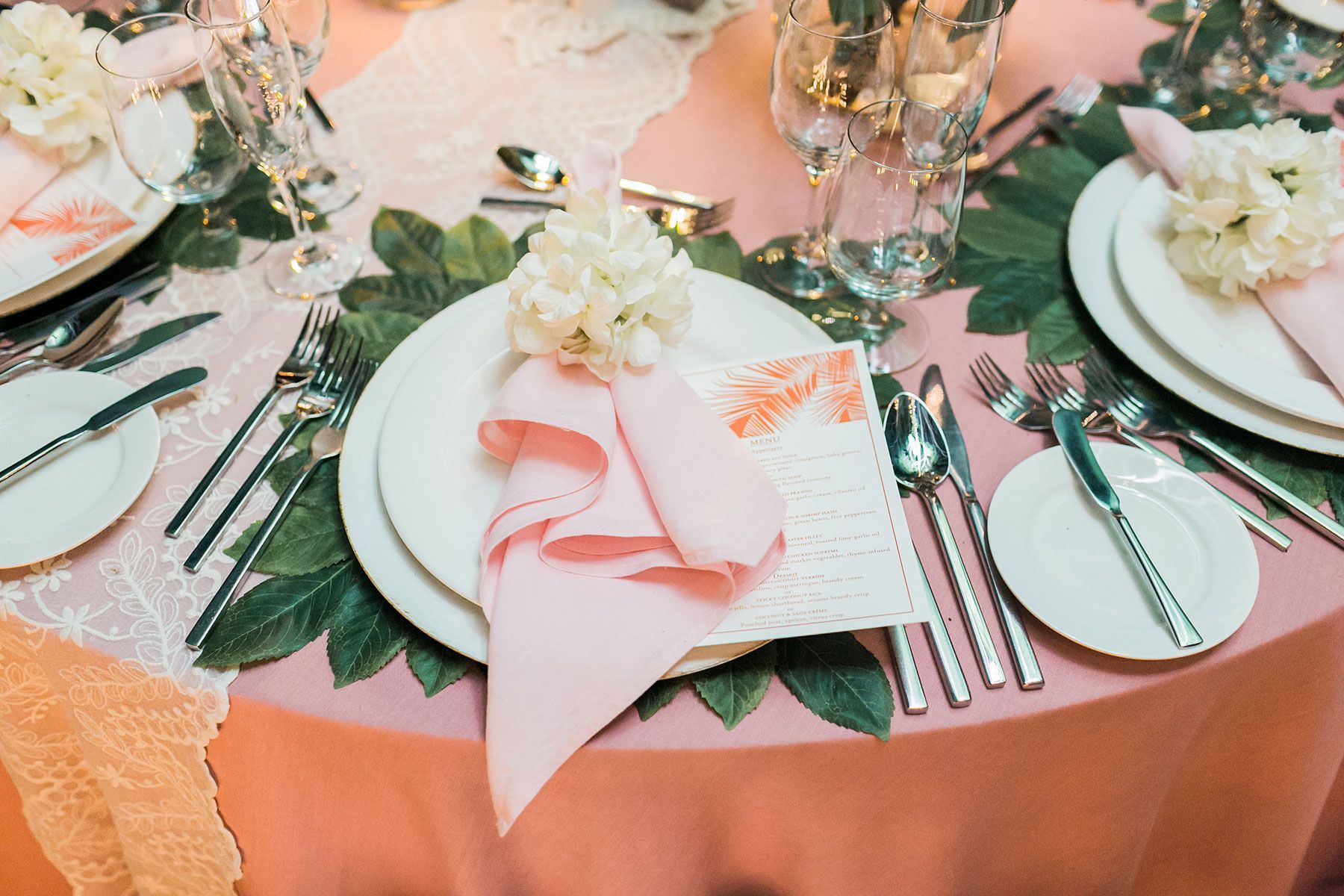 Elegant table setting with pink linen, white plates, and floral centerpiece, with green leaves as decoration.