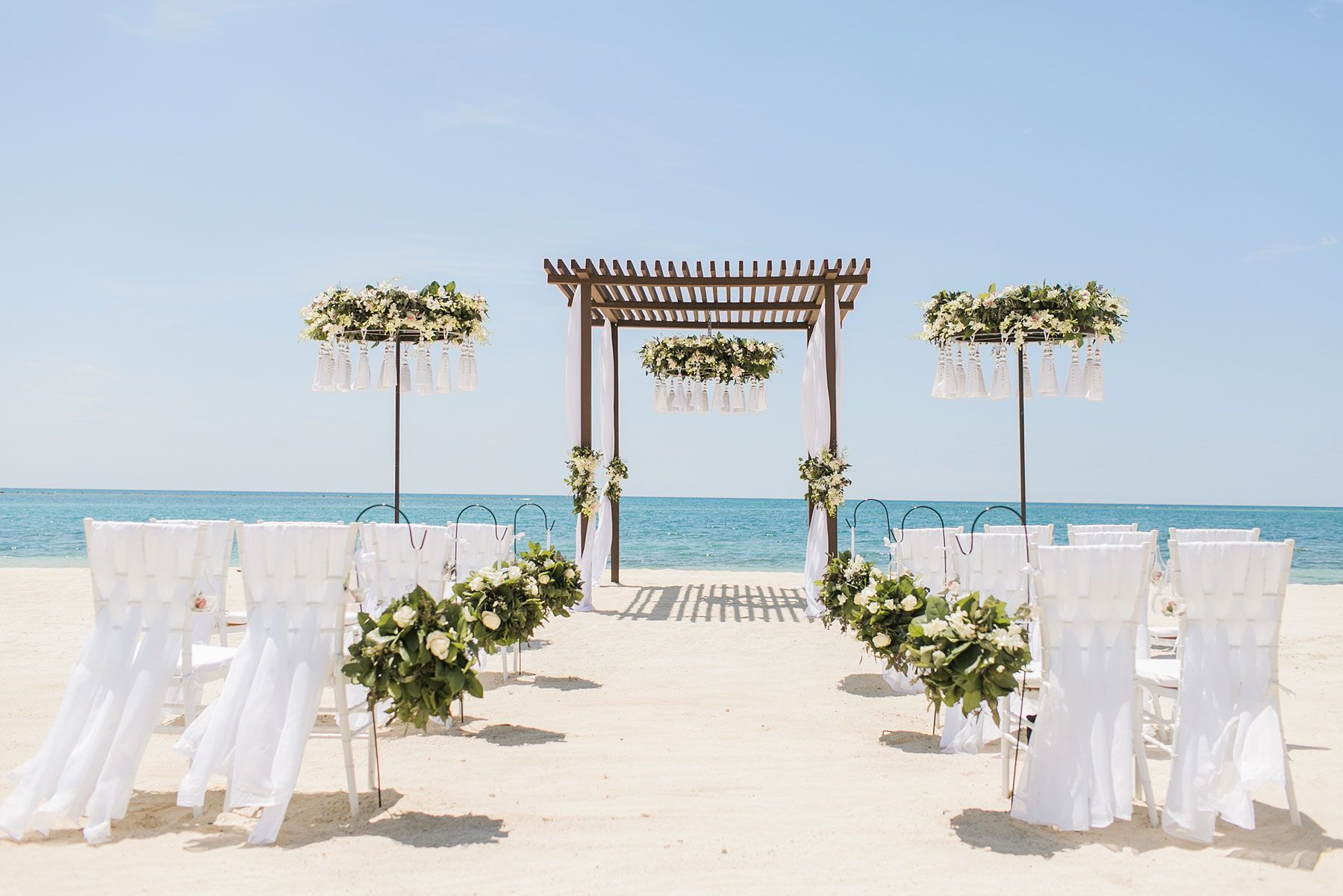 Beach wedding ceremony setup: white chairs, floral arrangements, wooden archway, ocean backdrop.