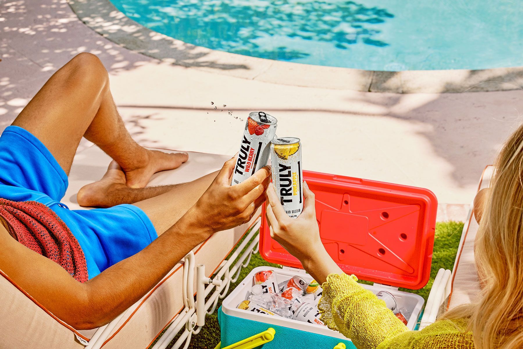 Two people toasting Truly hard seltzers by a pool. Cooler nearby.