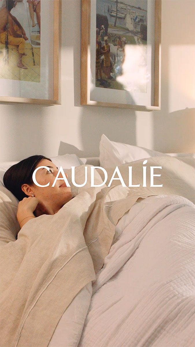 Woman in bed, smiling and relaxed, in a bright bedroom. Caudalie logo.