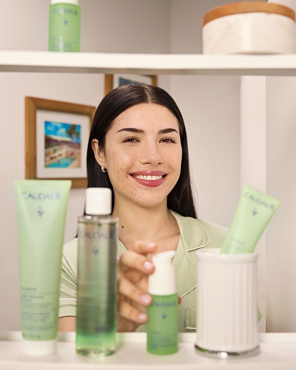 Woman smiling, reaching for a skincare product in a bathroom with green bottles on a shelf.