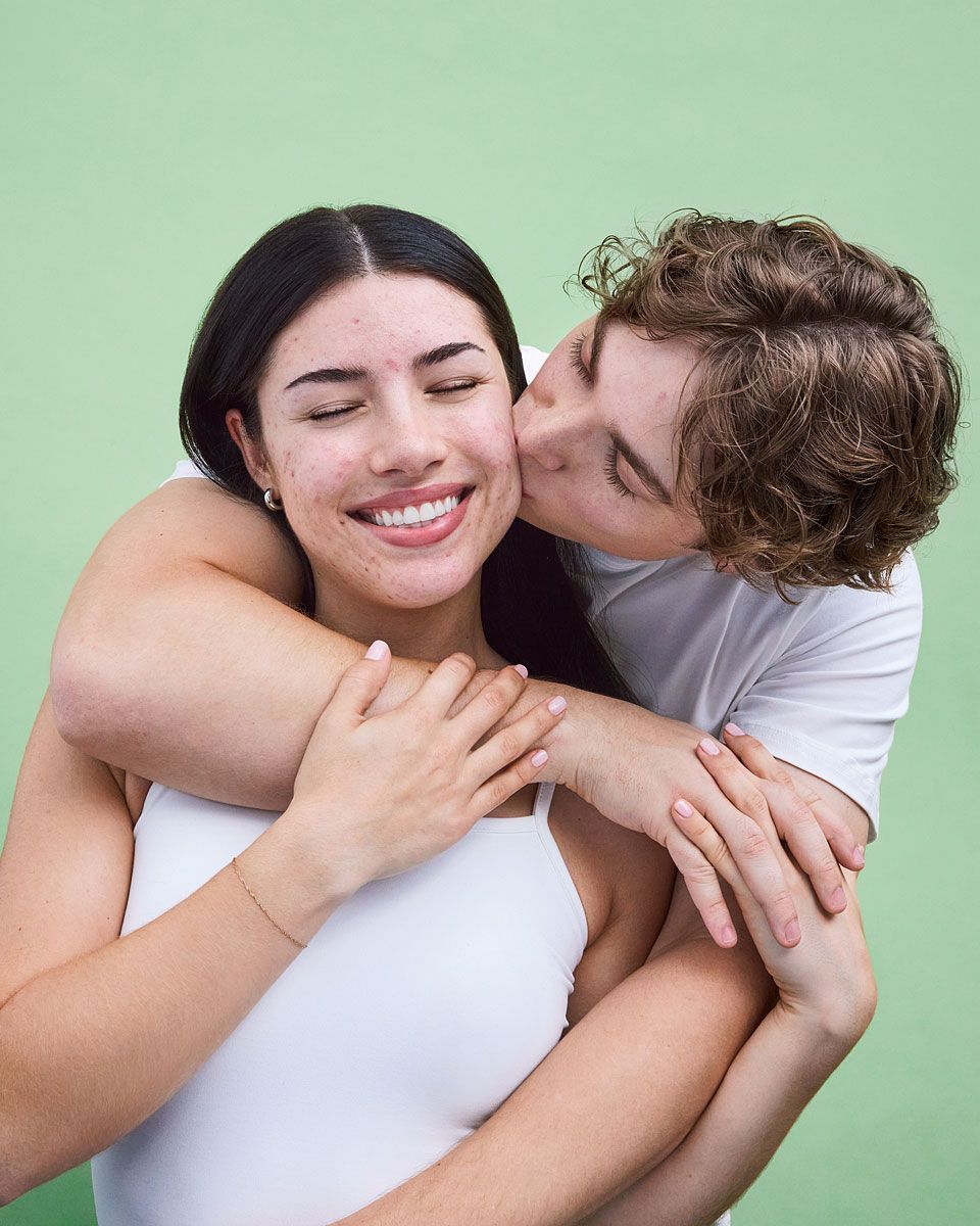 Man kissing woman on the cheek; woman smiling with arms around her. Green background.