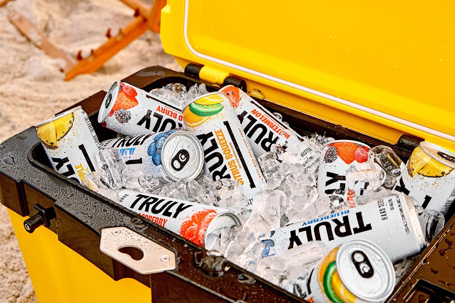 Yellow cooler filled with ice and cans of Truly hard seltzer on a sandy beach.