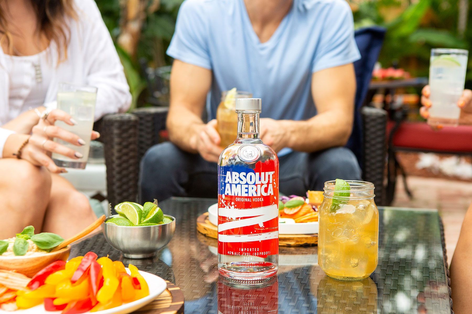 Absolut America vodka bottle on a table with drinks and people, outdoor setting.