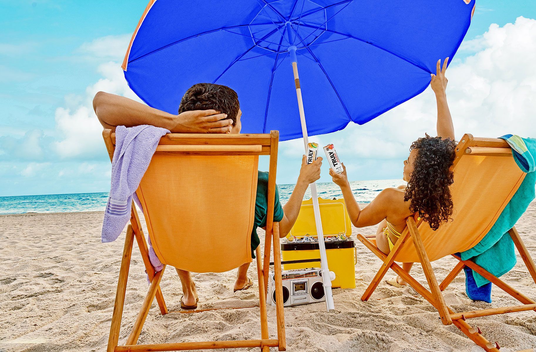 Two people relax in beach chairs under a blue umbrella, holding up drinks on a sandy beach.