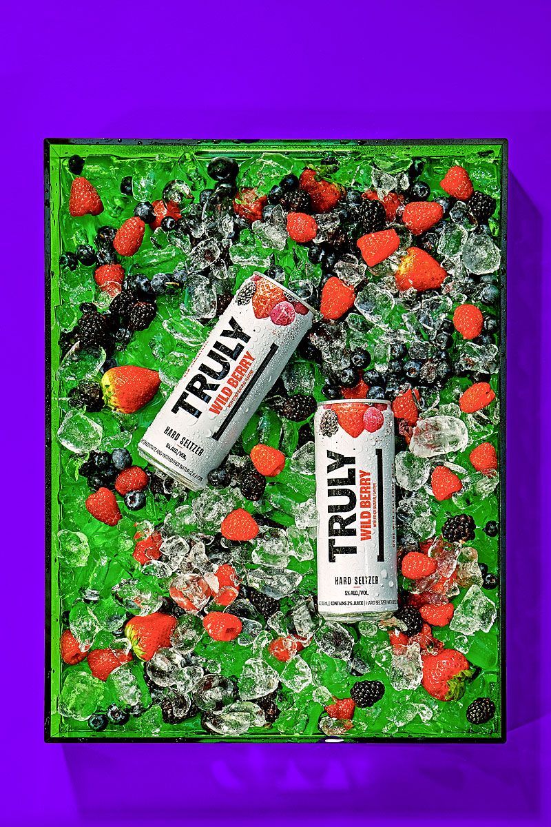 Two Truly Wild Berry hard seltzer cans surrounded by berries and ice on a green background.