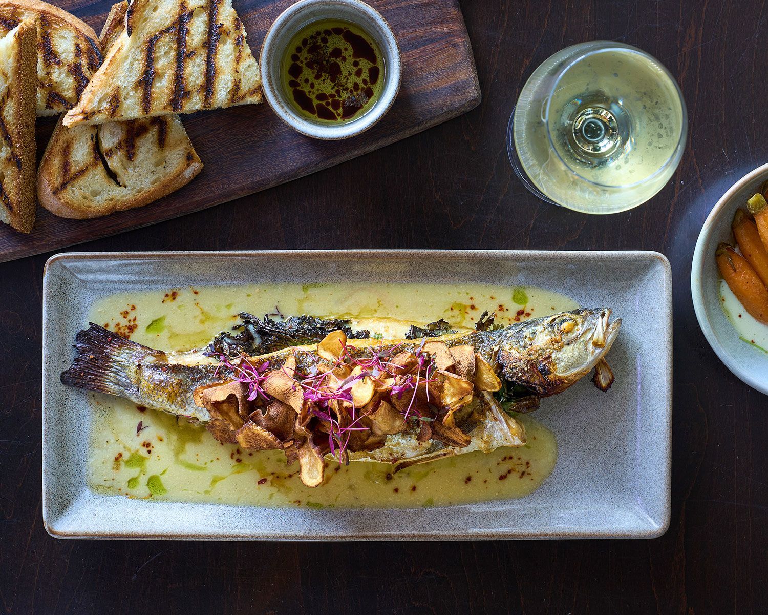Grilled whole fish on a plate with sauce, bread, dipping oil, wine, and sides on a dark wooden table.