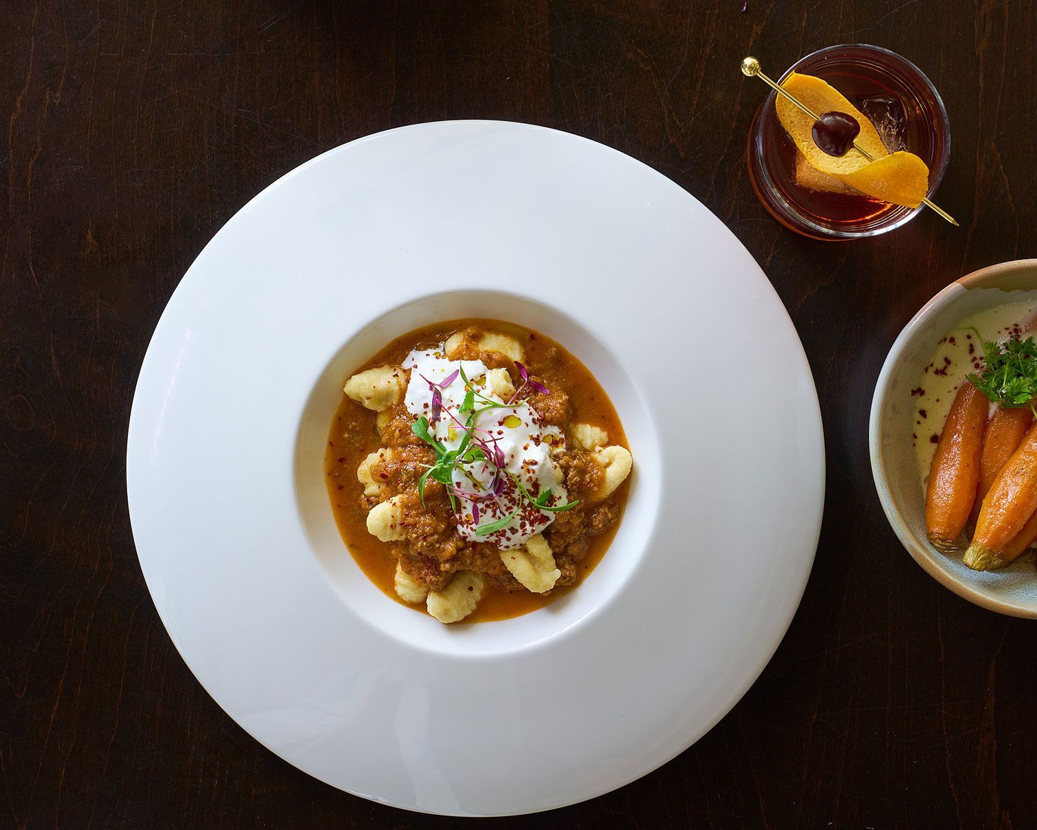Gnocchi in sauce topped with whipped cream, alongside a cocktail and carrots on a dark wooden table.
