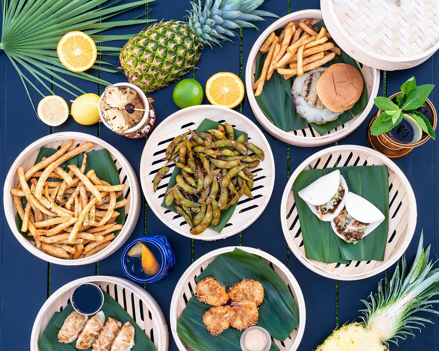 Overhead shot of various dishes: fries, dumplings, edamame, sliders, and fried chicken, arranged with fruit.