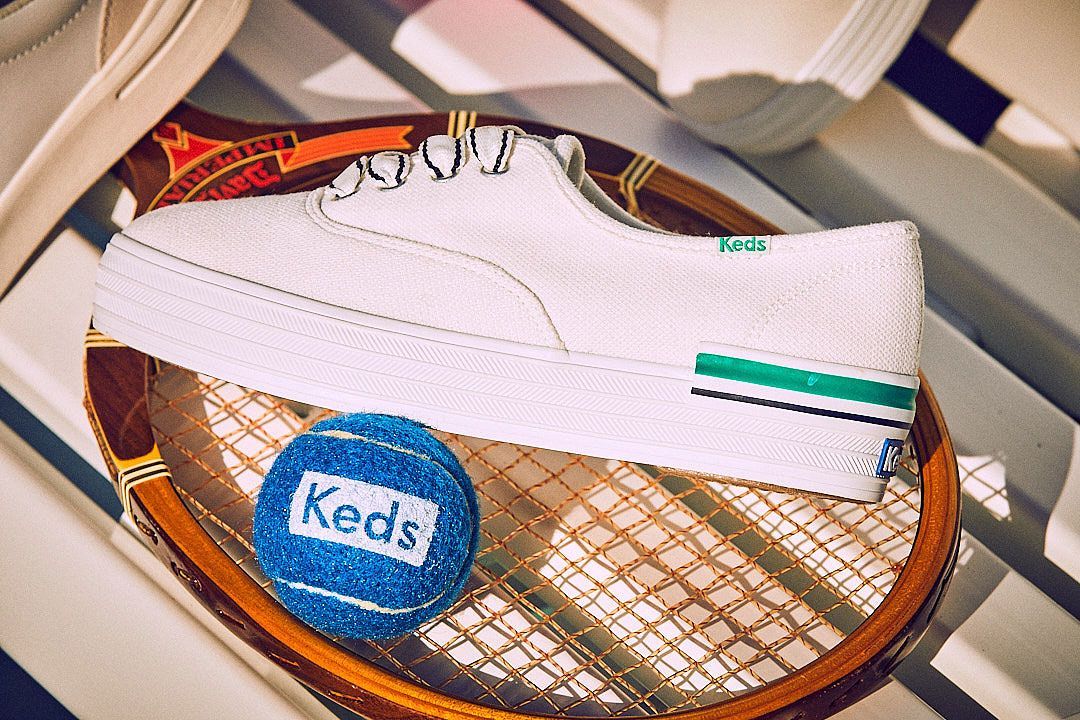 White Keds sneaker on a tennis racket, next to a blue Keds tennis ball.