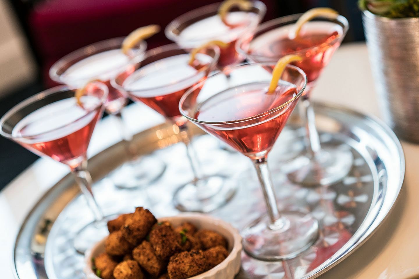 Cocktails in martini glasses with lemon twists on a silver tray, alongside a bowl of fried food.