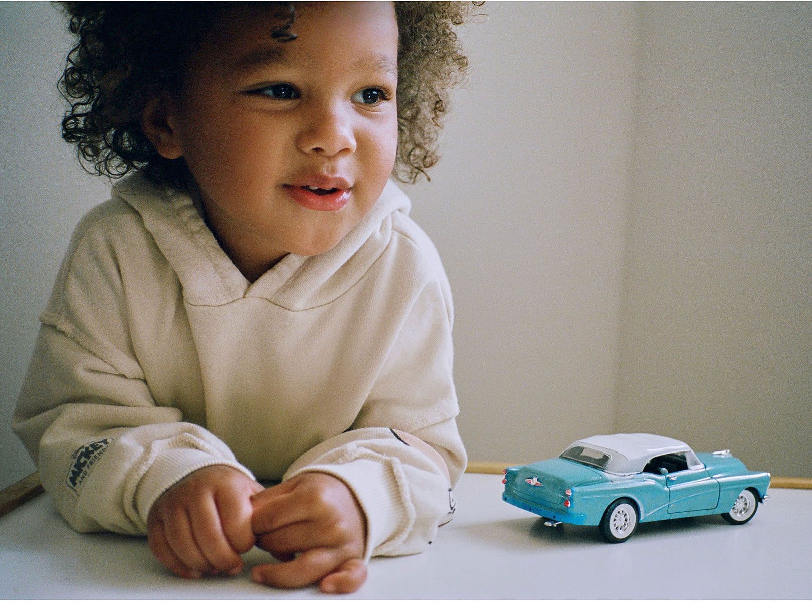 A child in a hoodie looks at a blue toy car on a white table.