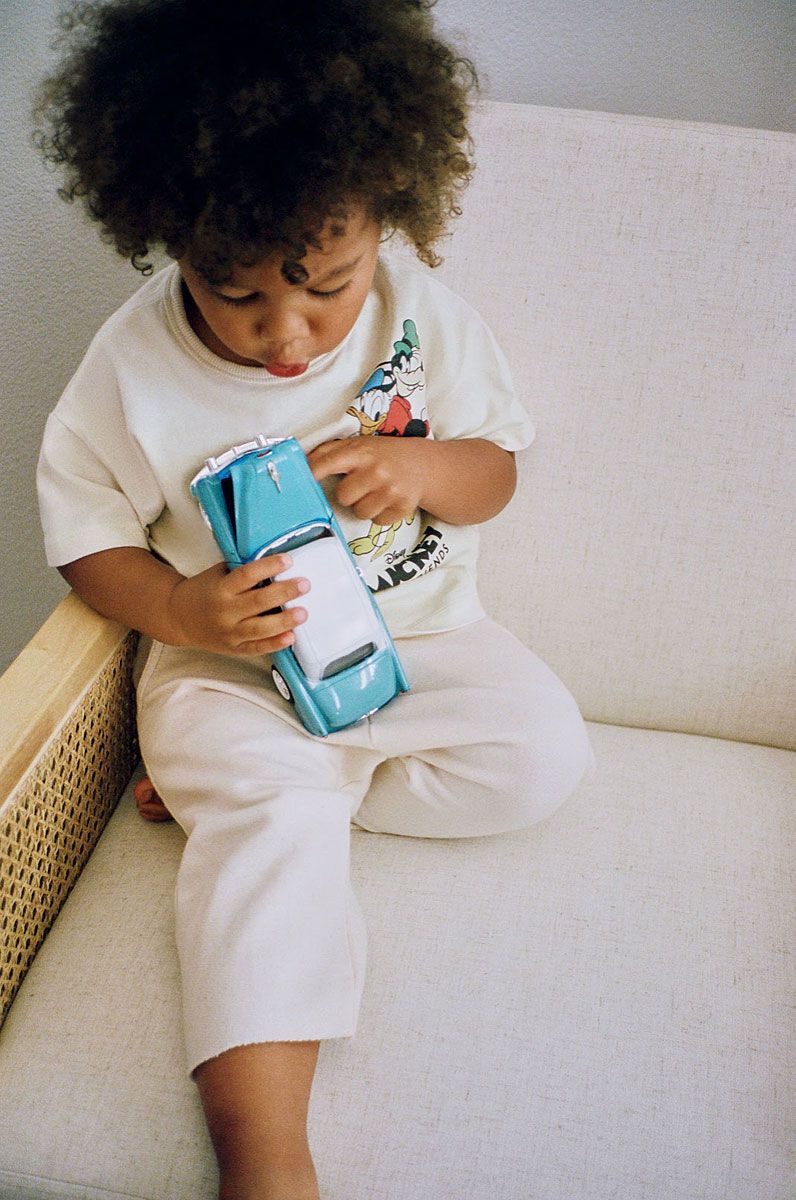 Child in cream-colored clothing sitting and holding a blue toy car.