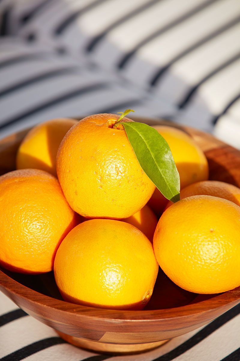 Oranges in a wooden bowl with a green leaf, on a striped fabric.