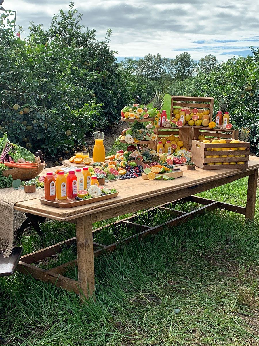 Long wooden table laden with fruits and juices, in an orchard setting.