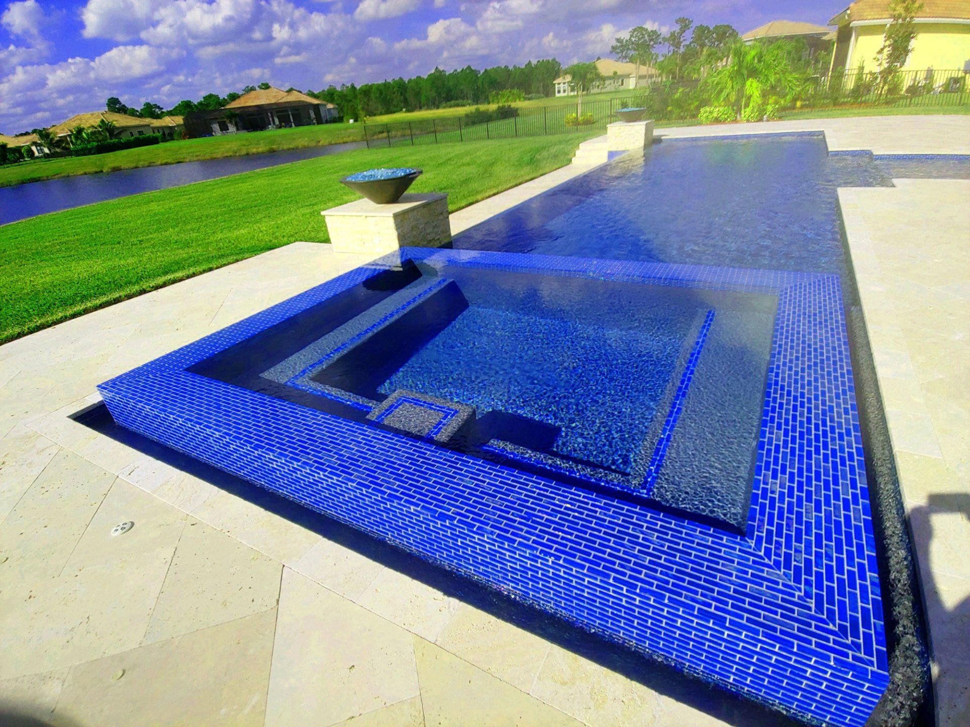 Blue tiled pool and spa, beige patio, green grass, lake, and houses under a cloudy sky.