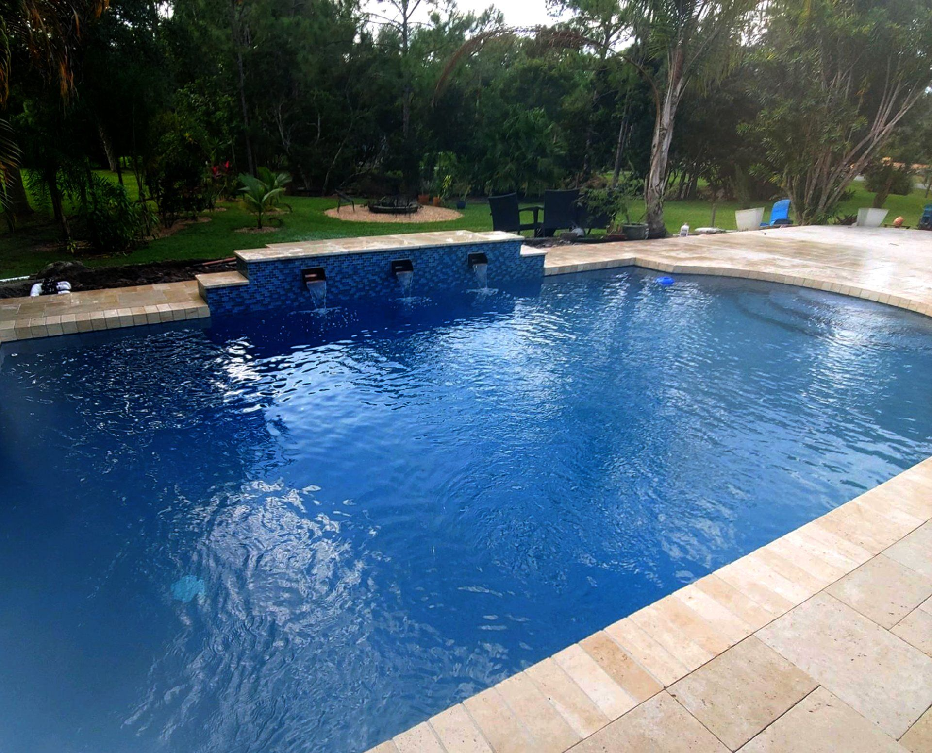 A blue tiled swimming pool with water fountains, surrounded by beige tiling. Trees in the background.